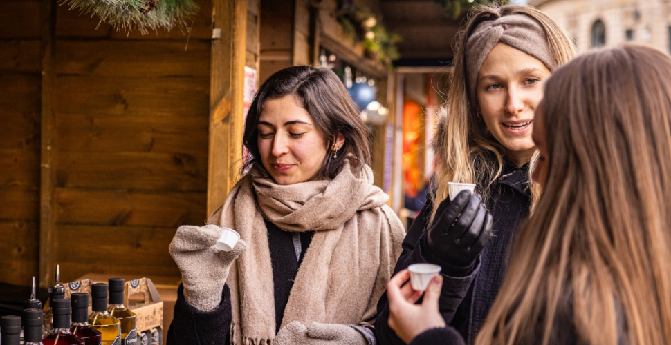 Three people dressed in winter clothing drinking festive beverages at an outdoor Christmas market stall with bottles on display.